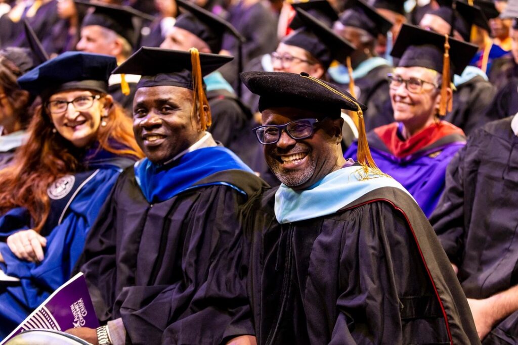 Faculty members wearing academic regalia with doctoral caps and blue hoods smiling together at a graduation ceremony.